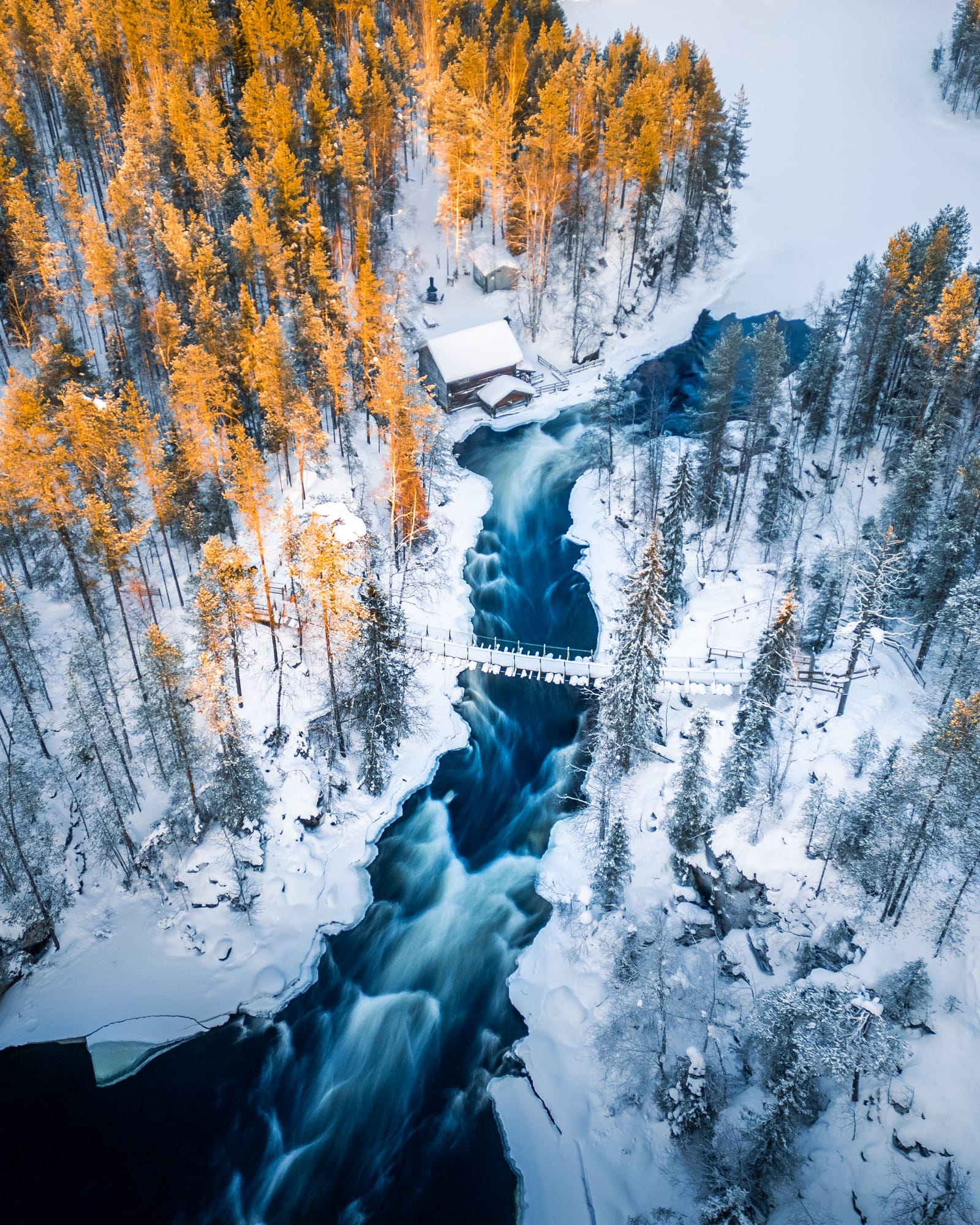 Aerial view of a dark river winding through snowy forest past a small cabin and footbridge.