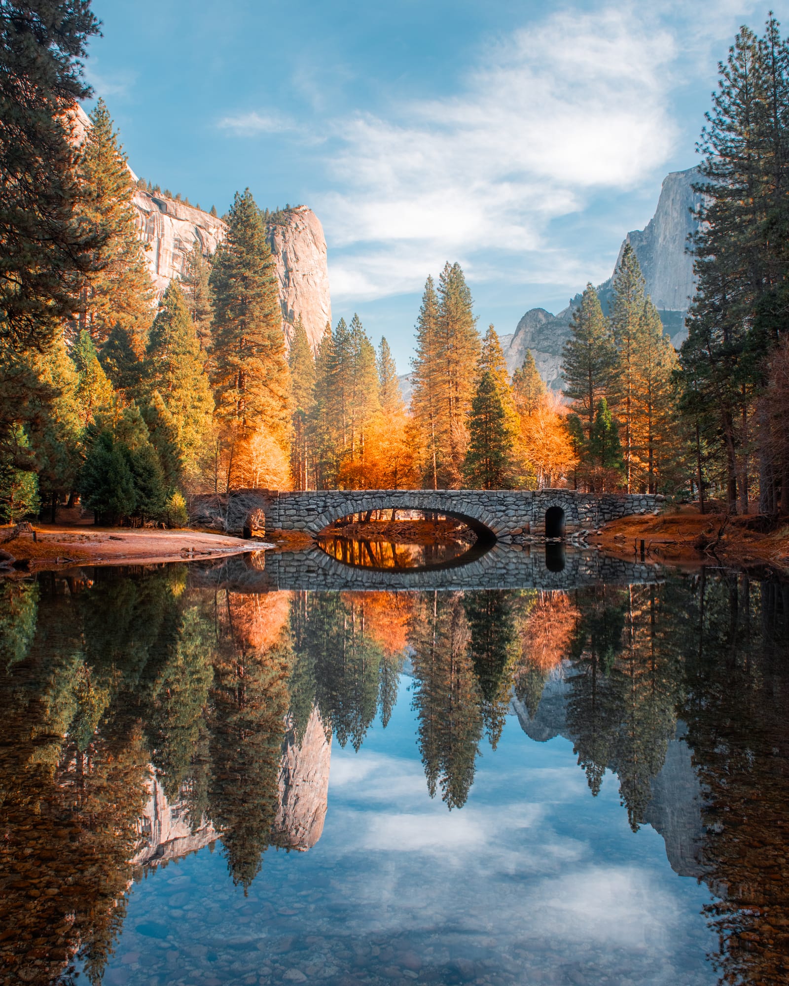 A stone bridge and golden autumn trees reflected in still water, with granite cliffs beyond.
