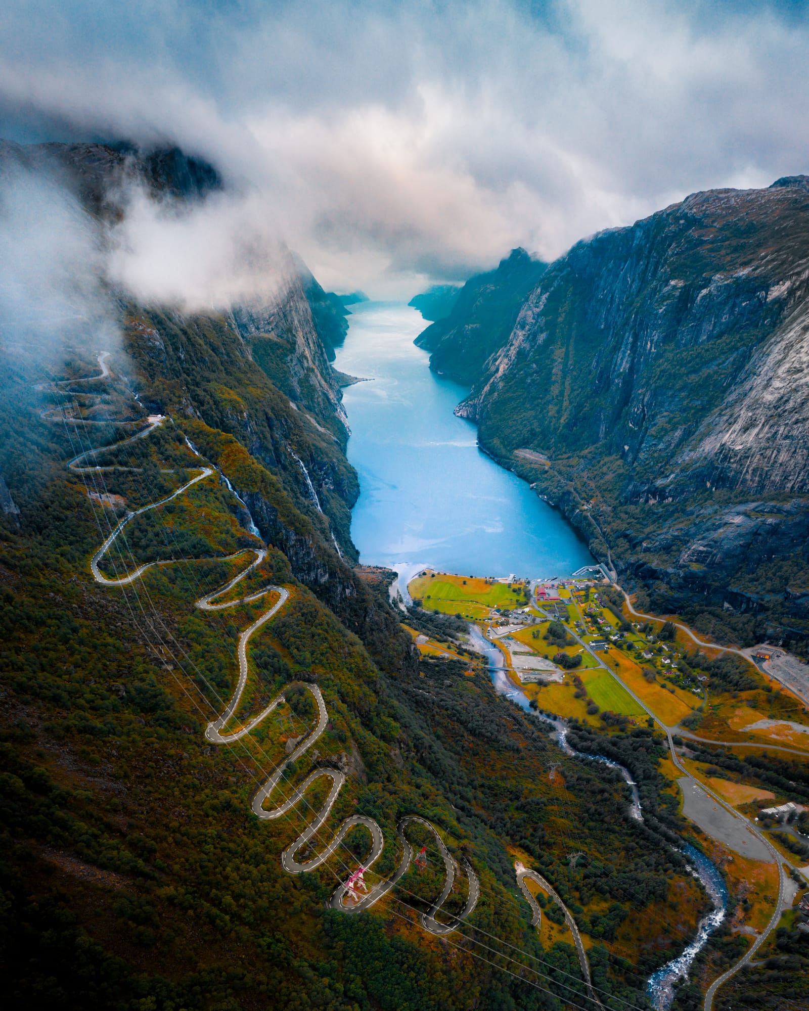 Aerial view of a switchback mountain road descending to a village at the head of a deep fjord under low clouds.