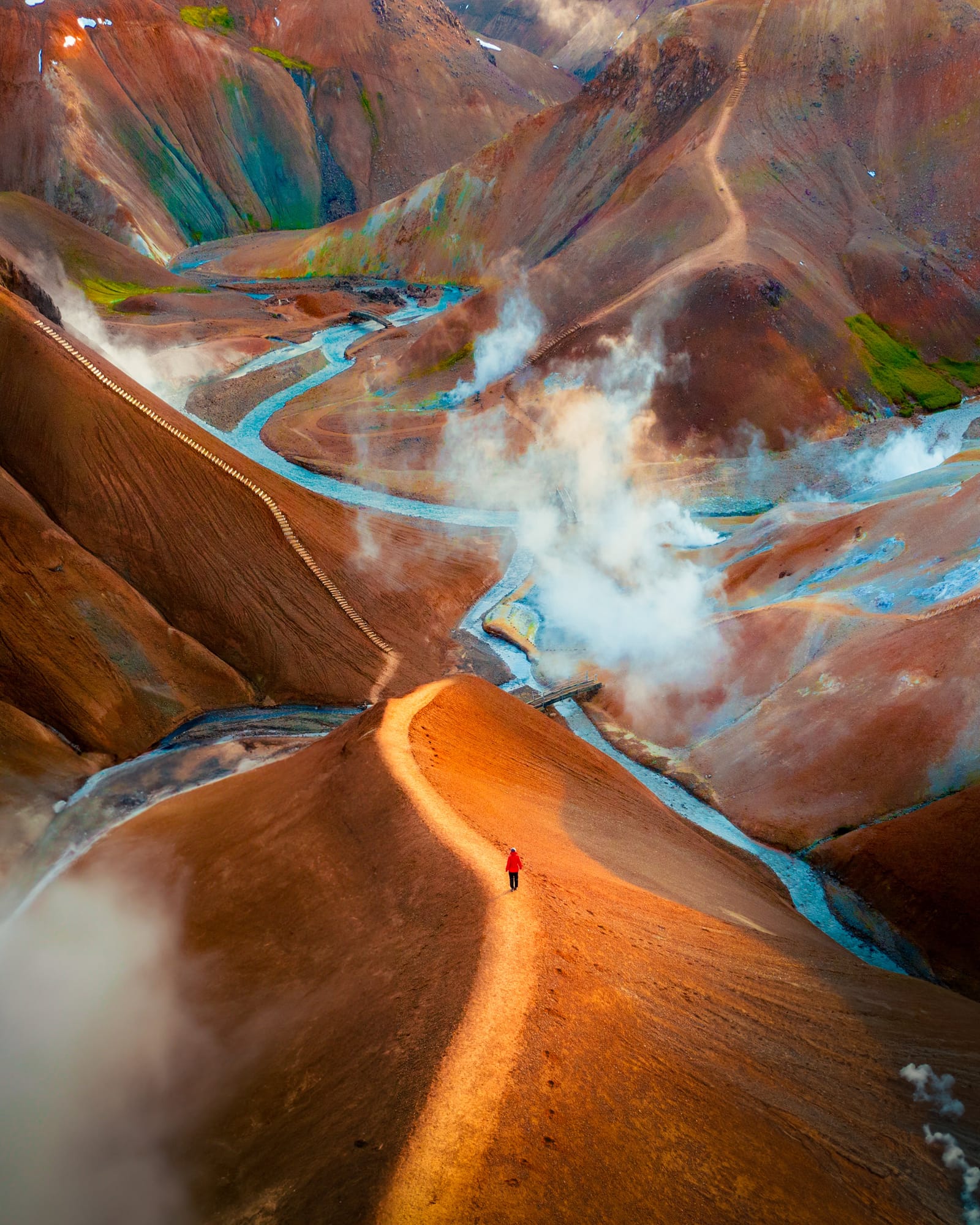 Aerial view of a hiker in red on a narrow ridge among orange and rust-colored geothermal hills, with steam and blue streams.