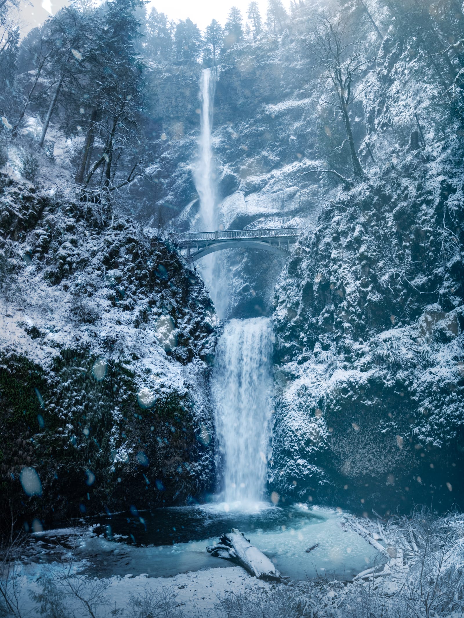 Multnomah Falls and its stone footbridge surrounded by snow-covered cliffs and trees.