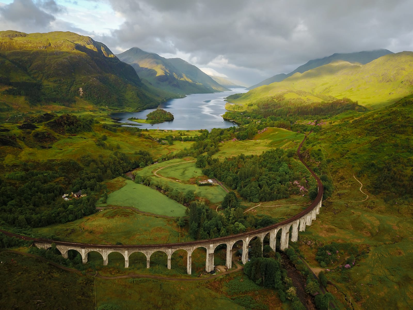 Aerial view of a curved stone railway viaduct crossing a green glen toward a long loch between mountains.