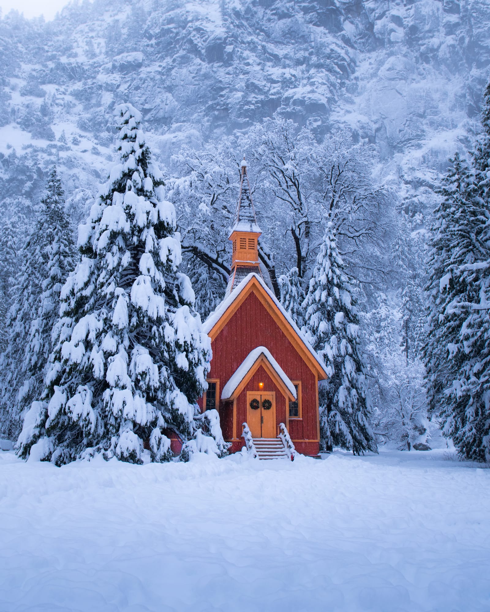 A small red chapel with a steeple among snow-laden pine trees after a snowfall.